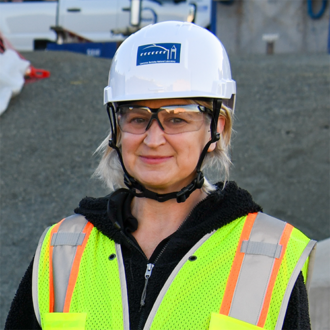 Portrait of a person with shoulder-length blonde hair wearing a hard hat, safety glasses, and safety vest, smiling.